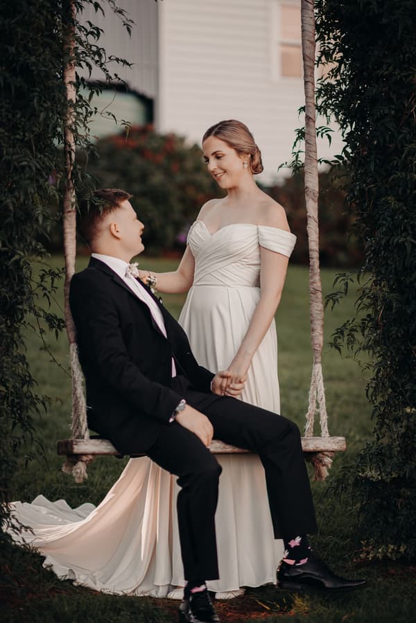Courtney the bride in an off-shoulder white wedding gown stands holding hands with Liam the groom, who is seated on a wooden swing framed by greenery at Tiffany's Maleny.
