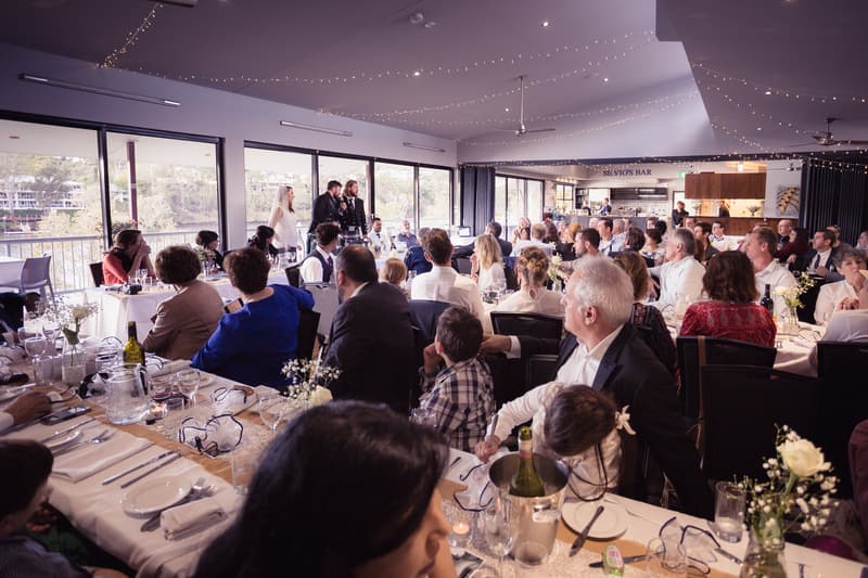 The bride Francesca and groom Ben stand together at the front of the Malouf Room at Toowong Rowing Club, addressing seated guests during the wedding reception.