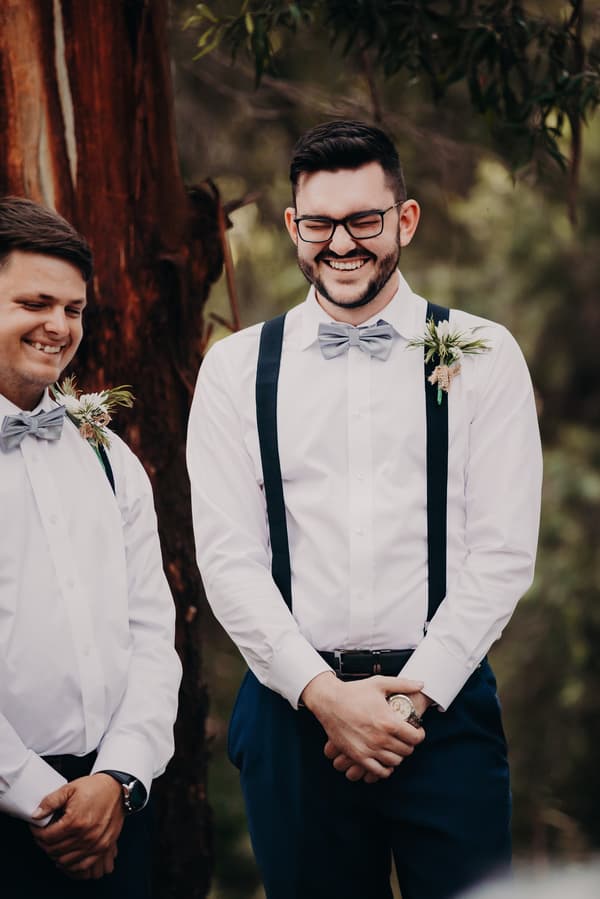The groom Trent and a groomsman stand outdoors near a tree at Ocean View Estates, both wearing white shirts with bow ties and suspenders, each with a boutonniere pinned to their shirts.