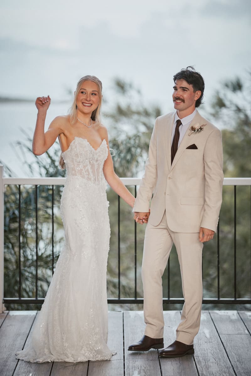 The bride Morgan and groom Joel stand holding hands on a wooden deck, with greenery and water visible in the background. Best time of year to get married by She Said Yes.