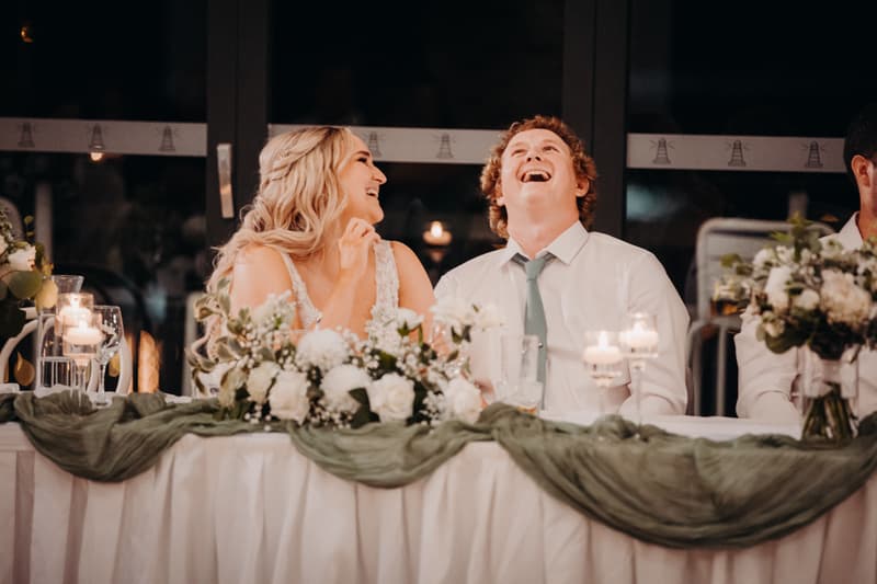 The bride Chloe and the groom Brodie sit at the head table decorated with white flowers and green fabric at Sandstone Point Hotel — Pumicestone Room during the reception stage, sharing a joyful moment.