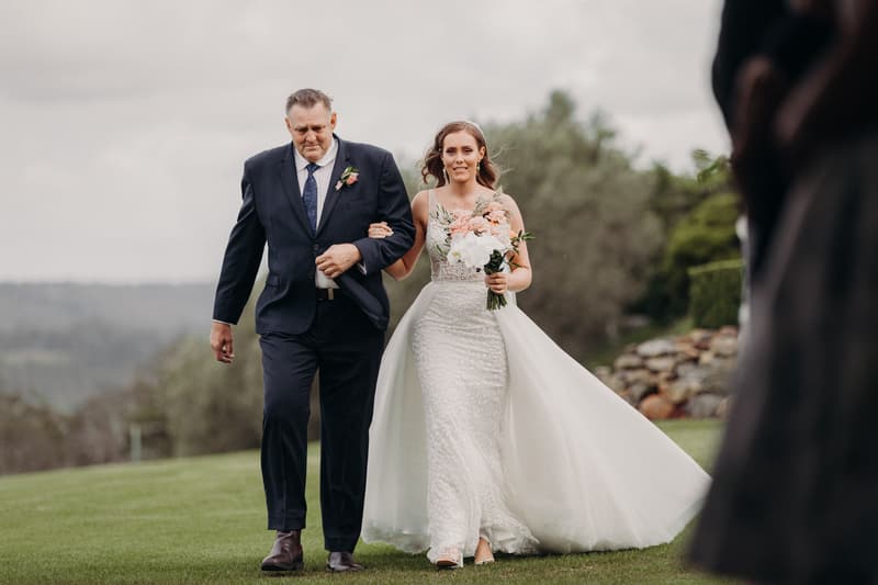 The bride Chelsea walks arm-in-arm with an older man, likely her father, outdoors on grass at Preston Peak Winery Functions Centre, holding a bouquet of flowers.