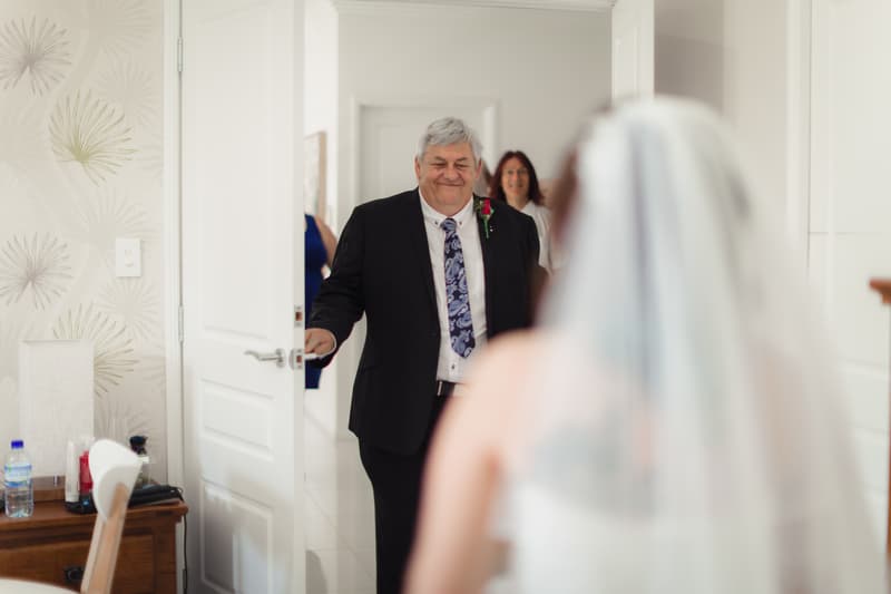 The bride stands facing the bride's father who is opening a door to enter the room, with a woman visible in the background.