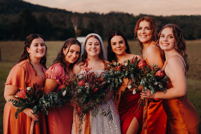 The bride Lilly poses with five bridesmaids outdoors at Yabbaloumba Retreat, all holding floral bouquets and smiling at the camera during the couple portraits stage.