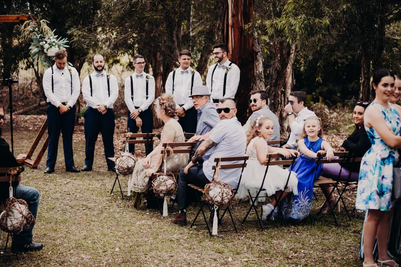 Five groomsmen stand near a wooden arch decorated with flowers at Ocean View Estates while guests, including children and adults, sit on folding chairs facing them outdoors.