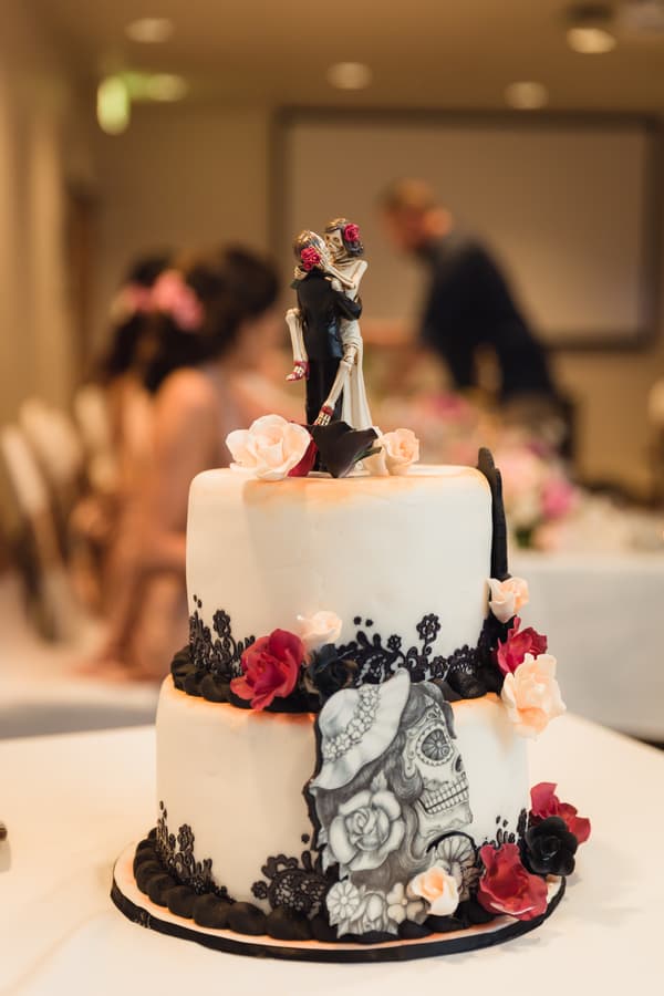 Two-tier wedding cake decorated with black lace patterns, red and white flowers, and a bride and groom skeleton figurine topper at Novotel Sunshine Coast Resort reception.