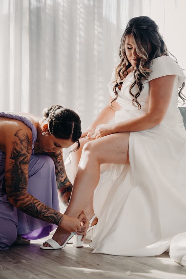 Tiffany, the bride in a white wedding dress, is seated while Brooke, wearing a lavender bridesmaid dress, helps her fasten a white high-heeled shoe at Sandstone Point Hotel — The Pavilion.