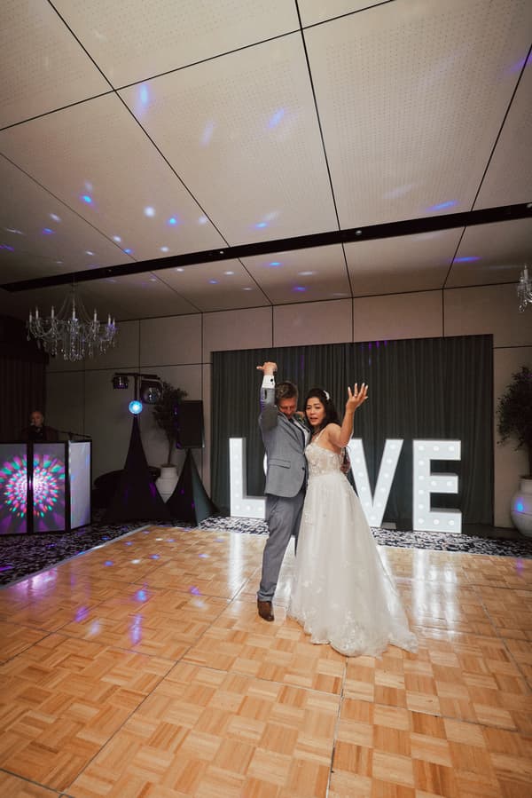 Bride Jheng and groom Dane dance on the parquet floor at the Sandstone Point Hotel — The Pumicestone Room reception, with illuminated LOVE letters in the background and a DJ booth to the side.