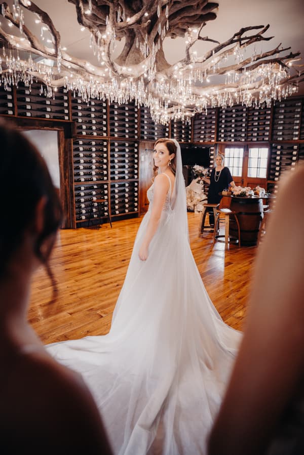 The bride Chelsea stands in a white wedding gown with a long train at Preston Peak Winery Functions Centre, looking back over her shoulder. Two blurred figures frame the foreground, and an older woman stands near a table with floral arrangements in the background.