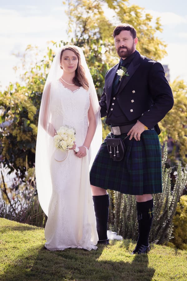 Bride Francesca in a white lace wedding gown holding a bouquet and groom Ben in a traditional Scottish kilt outfit pose outdoors with greenery in the background.