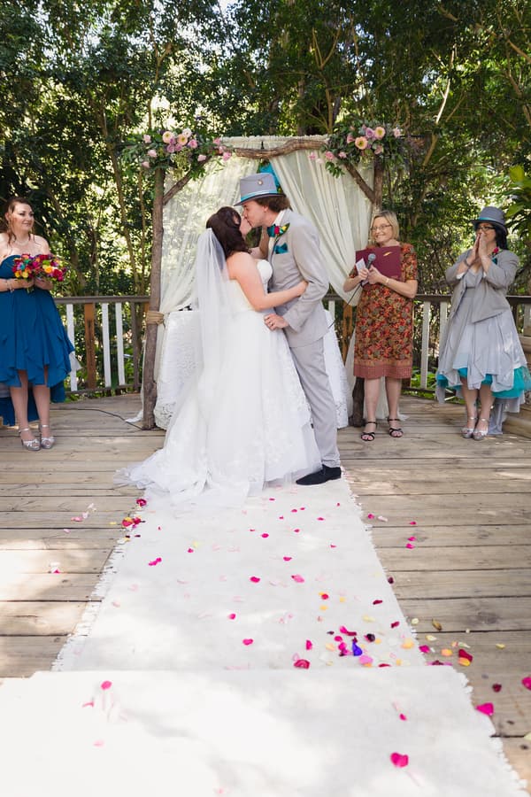 Bride Holly and groom Paul share a kiss at the altar during their wedding ceremony at Kwila Lodge, surrounded by a bridesmaid holding a bouquet, an officiant with a microphone and folder, and a guest clapping.
