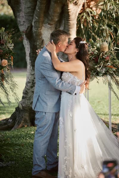 Bride Jheng and groom Dane share a kiss during their wedding ceremony, standing in front of a large tree with floral arrangements on either side. Destination wedding video packages by She Said Yes.