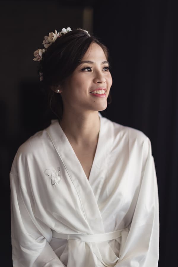 The bride Wing in a white satin robe with a floral hairpiece, smiling and looking to the side.