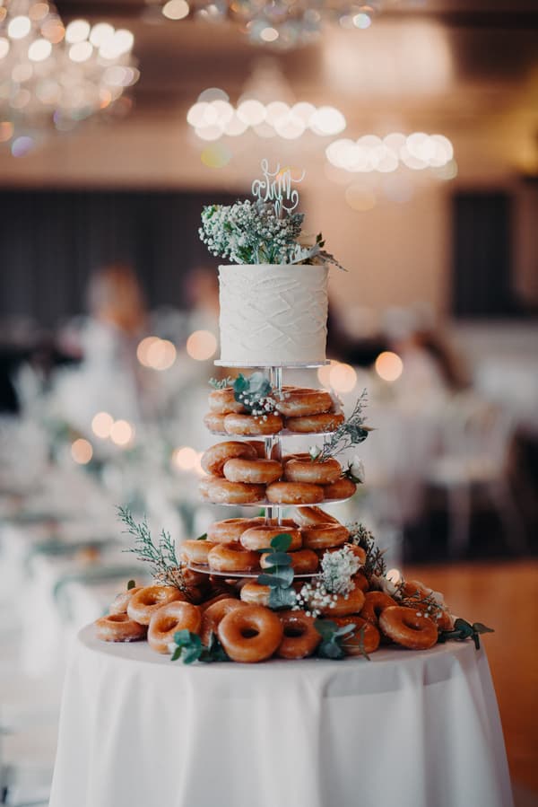 A wedding cake and donut tower decorated with flowers and greenery on a white tablecloth at Sandstone Point Hotel — Pumicestone Room reception.
