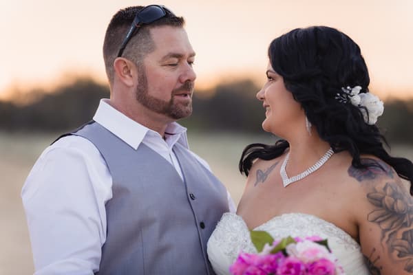 Kelly the bride and Shane the groom stand facing each other outdoors at sunset, with Kelly holding a bouquet of pink flowers and wearing a strapless wedding dress with visible tattoos and jewelry, and Shane wearing a white shirt and grey vest with sunglasses on his head. Wedding photographer Brisbane by She Said Yes.