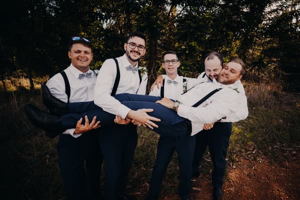 Four groomsmen dressed in white shirts, gray bow ties, and suspenders hold another groomsman horizontally outdoors with trees and grass in the background. Sunset wedding portraits Brisbane by She Said Yes.