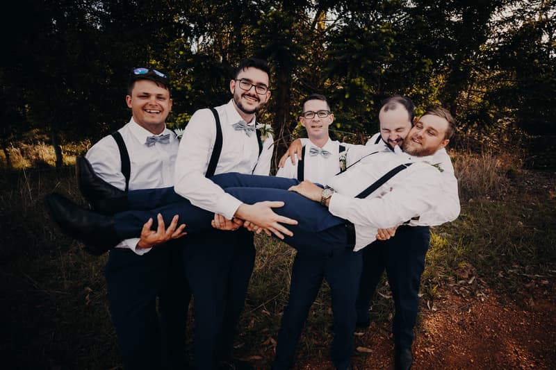 Four groomsmen dressed in white shirts, gray bow ties, and suspenders hold another groomsman horizontally outdoors with trees and grass in the background at Ocean View Estates.