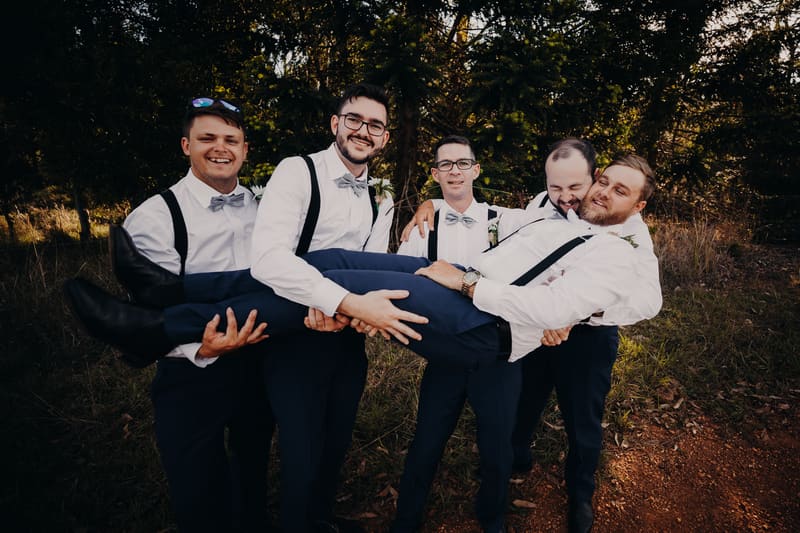 Four groomsmen dressed in white shirts, gray bow ties, and suspenders hold another groomsman horizontally outdoors with trees and grass in the background. Sunset wedding portraits Brisbane by She Said Yes.