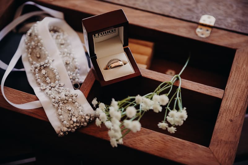 Wedding rings in a jewelry box alongside a beaded bridal sash and small white flowers on a wooden surface at Ocean View Estates.
