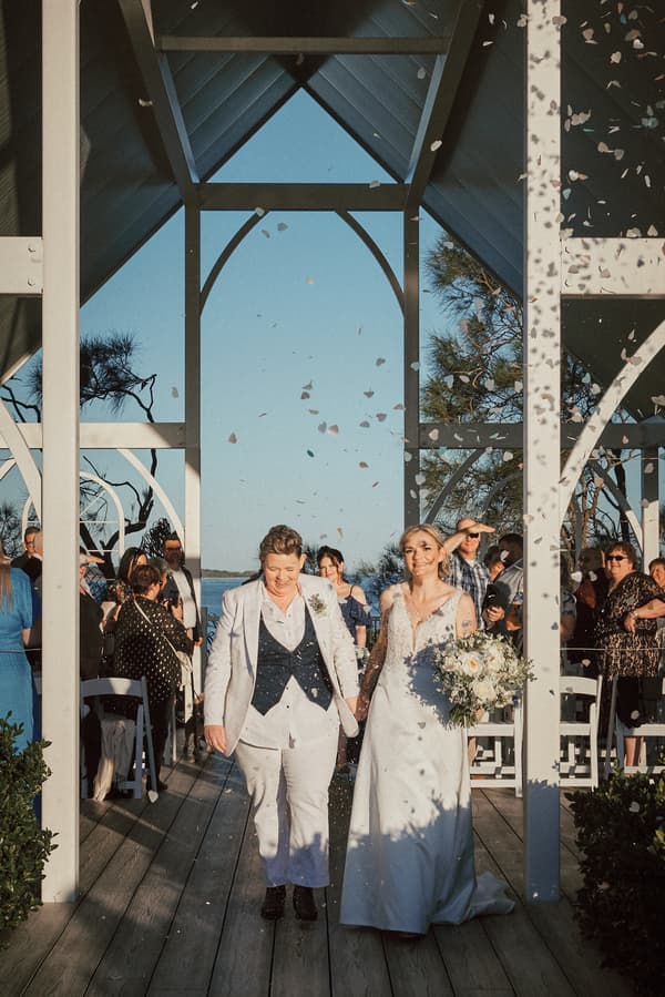 Brides Mandy and Selina walk down the aisle holding hands at the Sandstone Point Hotel — The Pavilion, with guests on either side and flower petals falling around them.