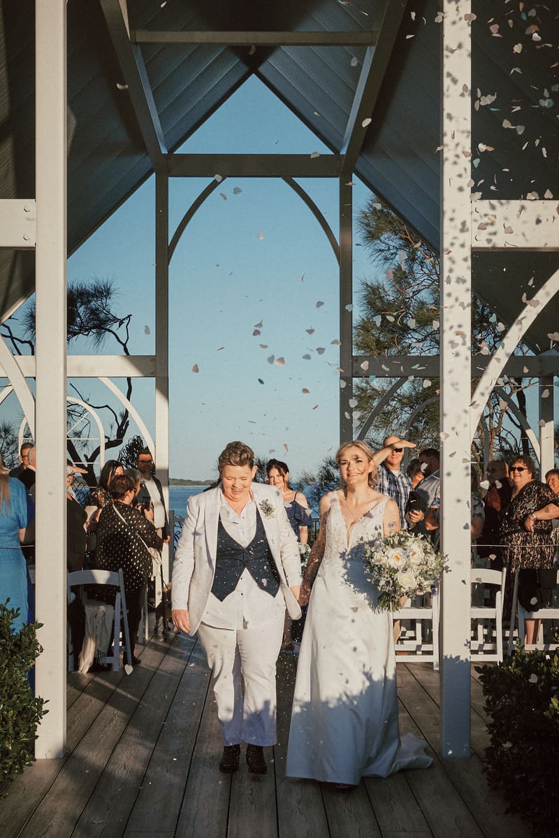Brides Mandy and Selina walk down the aisle holding hands at the, with guests on either side and flower petals falling around them. Brisbane videographer by She Said Yes.