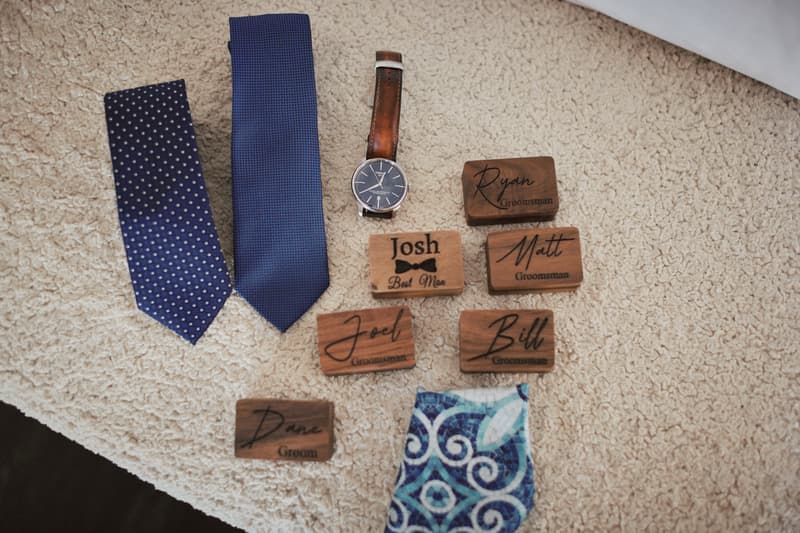 Two blue neckties, a wristwatch with a brown leather strap, a patterned blue pocket square, and wooden name tags for the groom Dane, best man Josh, and groomsmen Ryan, Matt, Joel, and Bill laid out on a beige textured surface.