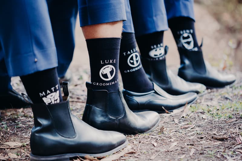 Close-up of four groomsmen's black dress shoes and personalized black socks with names and 'Groomsman' text, standing on a natural ground surface.