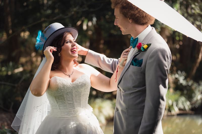 The bride Holly wearing a white wedding dress and a grey hat with blue feathers adjusts her hat while smiling at the groom Paul, who is dressed in a light grey suit with a blue bow tie and colorful boutonniere, outdoors at Kwila Lodge.
