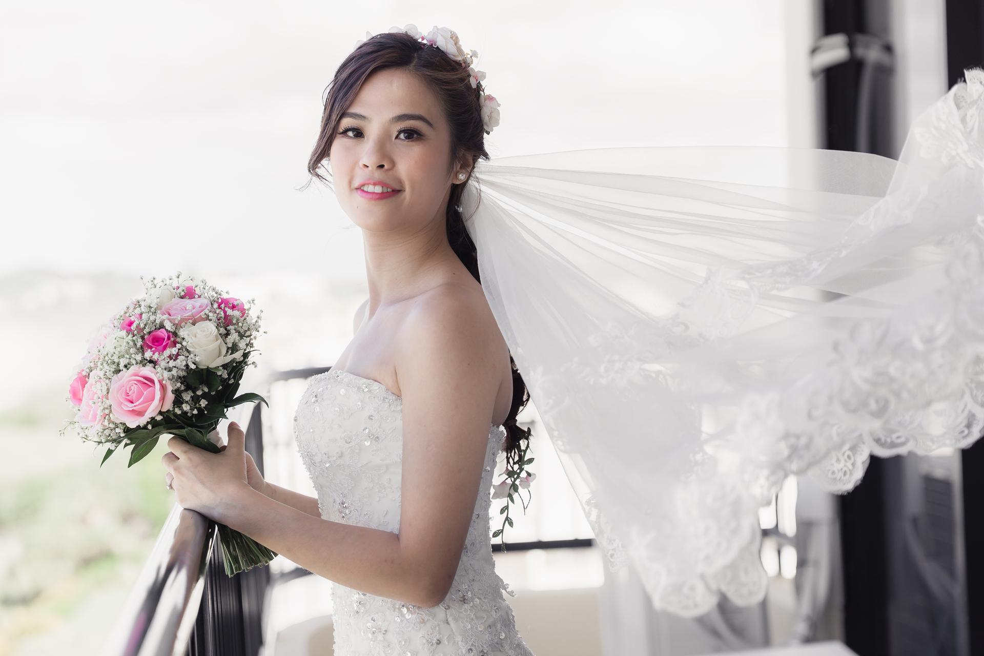 The bride Wing stands holding a bouquet of pink and white flowers with her veil flowing behind her at Bilinga Beach Weddings.