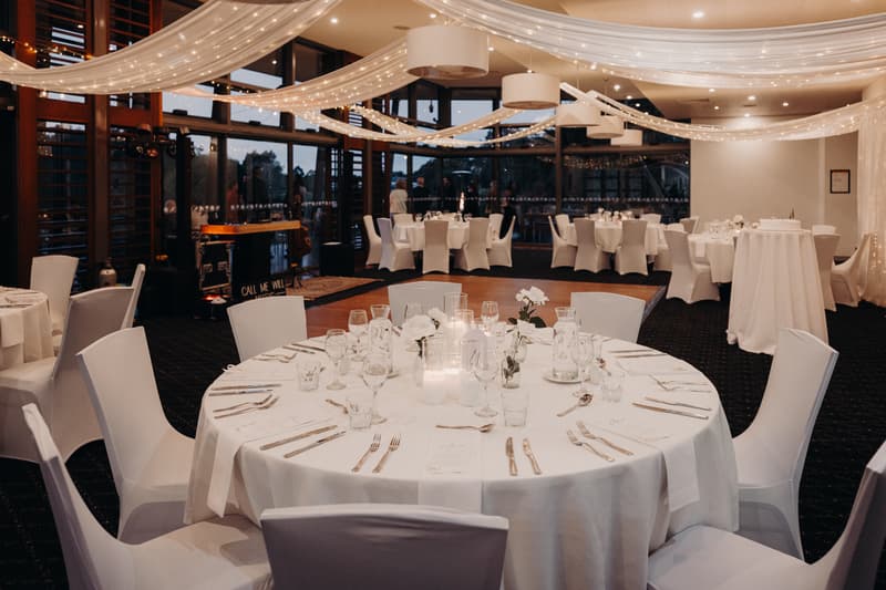Reception tables set with white linens, glassware, cutlery, and floral centerpieces in The Pandanus Room at The Tides, decorated with draped fabric and string lights overhead.