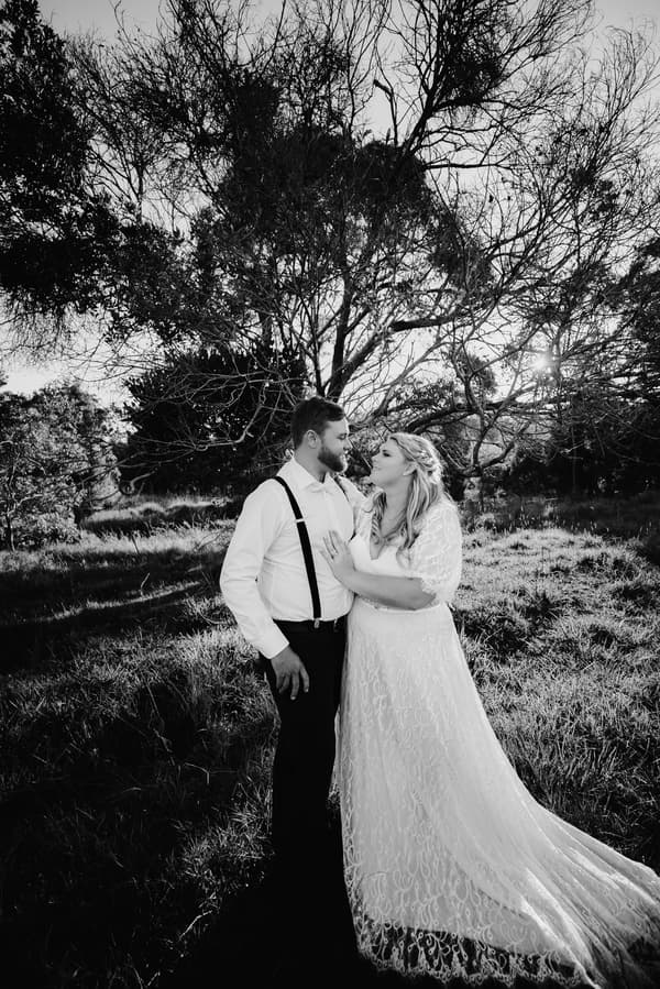 Annie in her wedding dress and Trent in a white shirt with suspenders stand close together outdoors at Ocean View Estates, with trees and grass in the background.