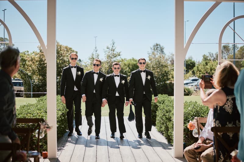 Four groomsmen in black tuxedos and sunglasses walk down the aisle at Sandstone Point Hotel — The Pavilion, with guests seated and watching.