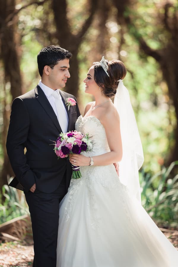 Pasha and Maryam pose together outdoors in a scenic setting, with Maryam holding a bouquet of purple and pink flowers and wearing a tiara and veil.