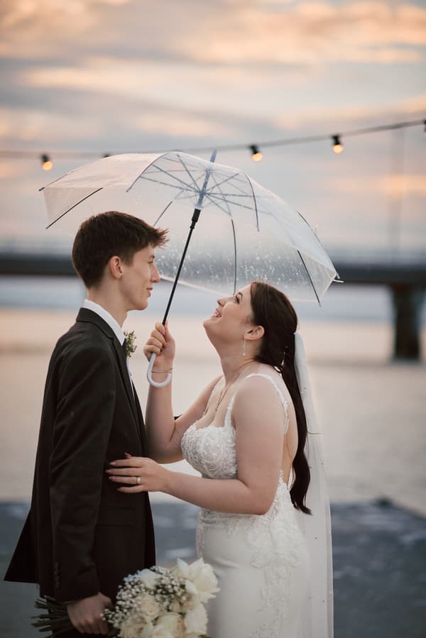 Terion the bride and Tyler the groom stand close under a clear umbrella at Sandstone Point Hotel, with the bride holding the umbrella and the groom holding a bouquet of white flowers.