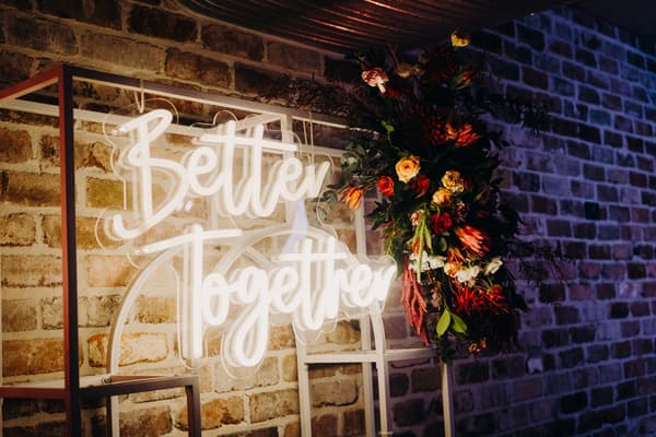 Neon sign reading 'Better Together' mounted on a white frame with a floral arrangement on the right, set against a sandstone brick wall at Sandstone Point Hotel — The Cellar.