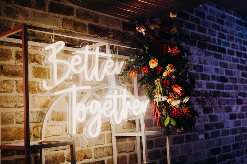 Neon sign reading 'Better Together' mounted on a white frame with a floral arrangement on the right, set against a sandstone brick wall at Sandstone Point Hotel — The Cellar.