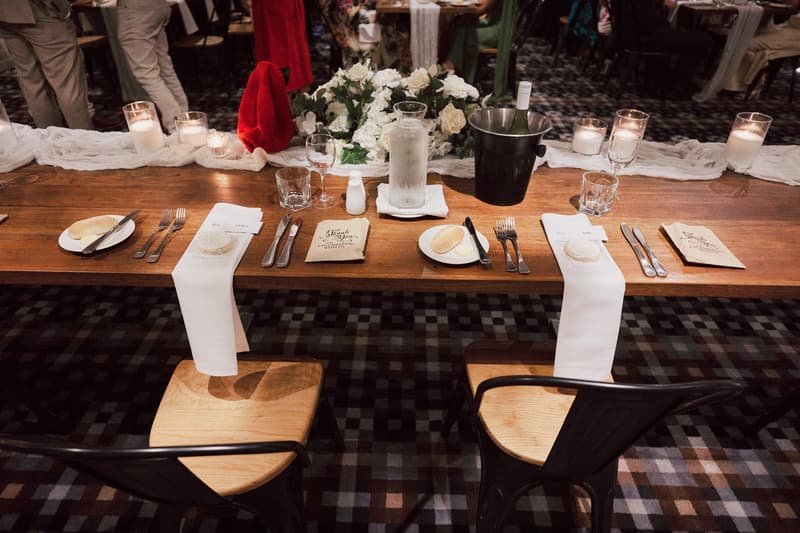 Reception table set for bride and groom at Sandstone Point Hotel — The Cellar, featuring white napkins, plates with bread rolls and butter, cutlery, glassware, candles, a floral centerpiece, and a wine bottle in an ice bucket.
