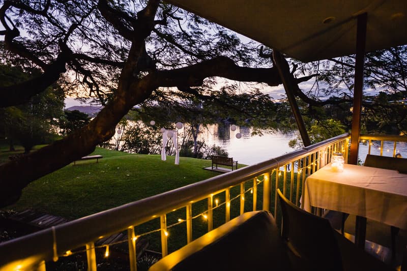 View from the balcony of The Malouf Room at Toowong Rowing Club overlooking a grassy area with a wedding arch decorated with white fabric and hanging lanterns near the water at sunset.