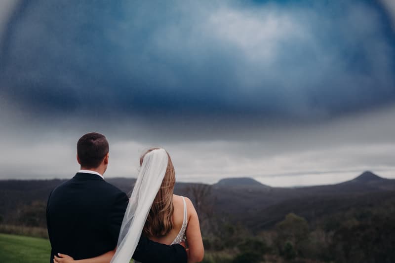 Bride Chelsea and groom Brent stand with their backs to the camera, overlooking a scenic mountainous landscape at Preston Peak Winery Functions Centre.