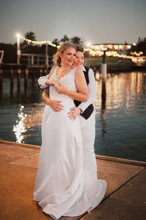Mandy and Selina embrace near the water at Sandstone Point Hotel during a styled couple portrait session at dusk with string lights in the background.