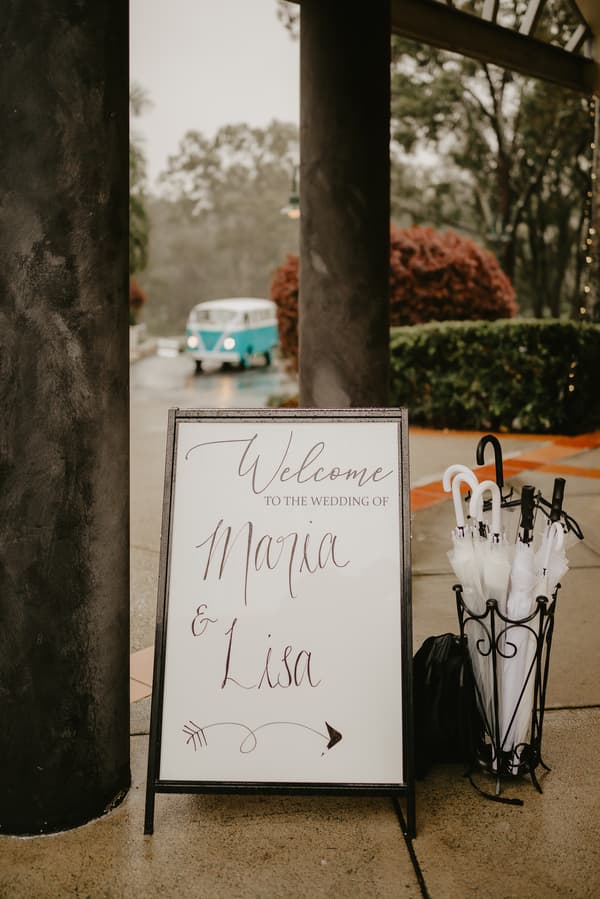 A welcome sign for the wedding of Maria and Lisa is displayed outdoors next to a stand holding several umbrellas, with a vintage blue and white van visible in the background.