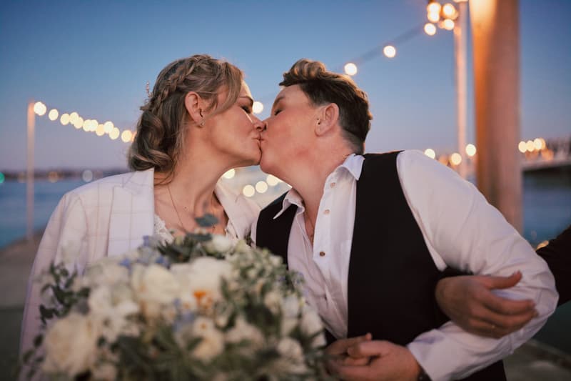 Mandy and Selina share a kiss outdoors at Sandstone Point Hotel during their couple portraits session, with string lights and water in the background and Mandy holding a bouquet of white flowers.