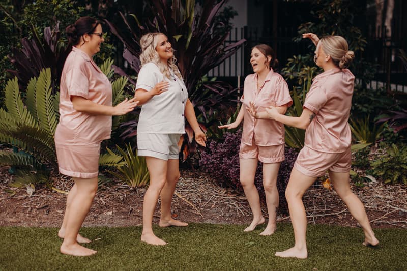 The bride Chloe in white pajamas and three bridesmaids in matching pink pajamas stand barefoot on grass outside, interacting and smiling near garden plants at Sandstone Point Hotel.