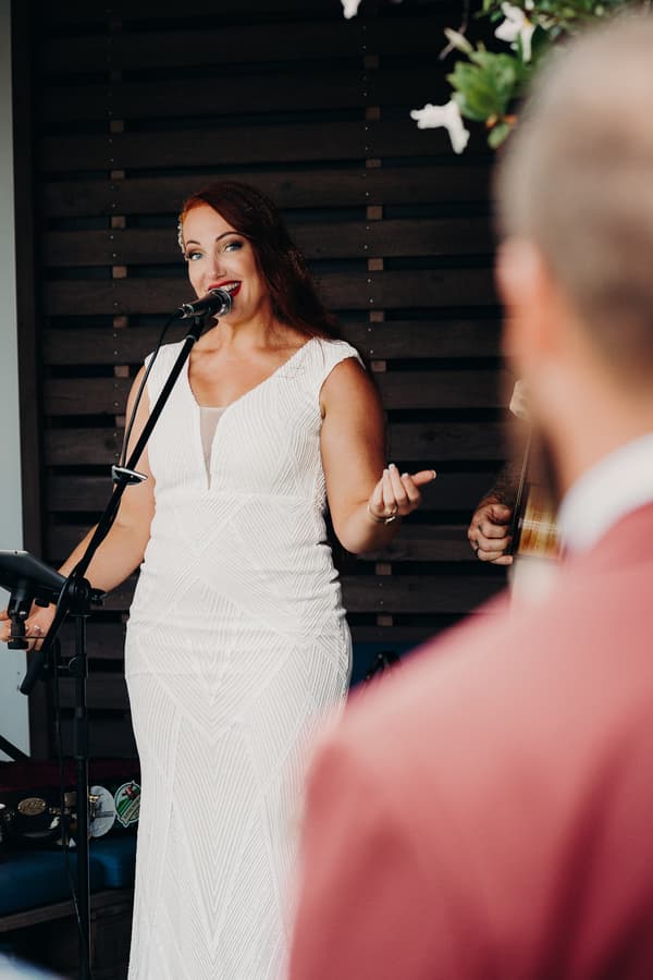 The bride in a white wedding dress speaks or sings into a microphone at the reception stage, with the groom partially visible in the foreground at Sandstone Point Hotel — The Cellar.