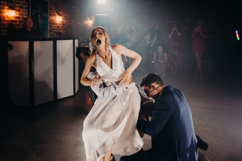 Courtney, the bride, is laughing with her mouth wide open while Cameron, the groom, kneels in front of her lifting her white wedding dress at the Sandstone Point Hotel — Cellar during the reception stage. Guests are visible in the background watching and taking photos, and a man stands behind a DJ booth on the left.