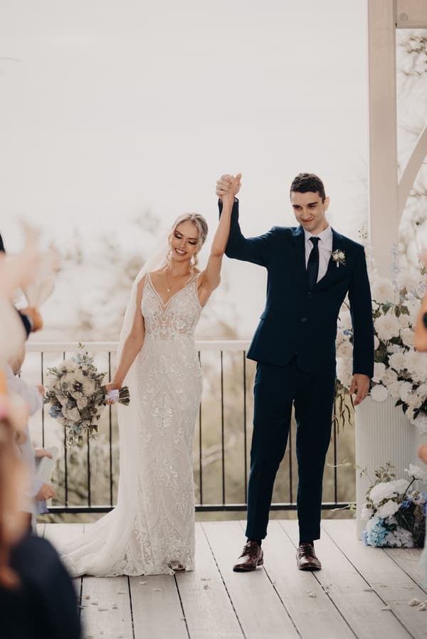 Courtney and Cameron stand holding hands raised together at the Sandstone Point Hotel — Pavilion ceremony stage, with Courtney holding a bouquet and guests applauding around them.
