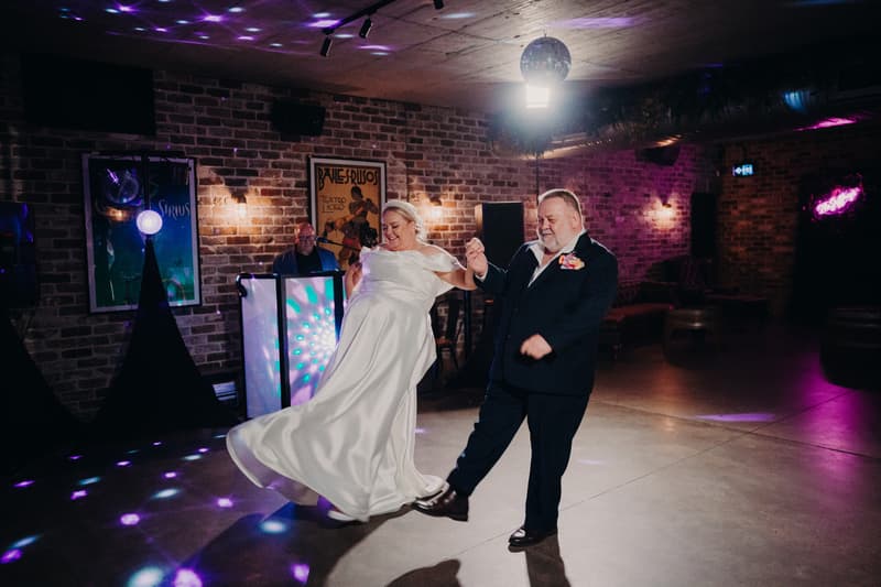 The bride Jacquelyne dances with an older man, likely her father, on the dance floor at Sandstone Point Hotel — Cellar during the wedding reception. A DJ is visible in the background with colorful lights and brick walls surrounding the space.