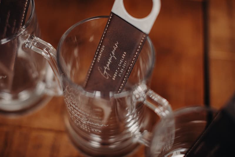 A close-up of a leather bottle opener inside a clear glass mug on a wooden surface, the bottle opener engraved with the names Brent and Chelsea and the date 21.03.2021.