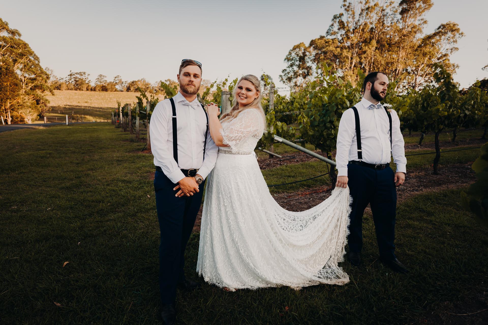 The bride Annie stands between two groomsmen at Ocean View Estates, with one groomsman holding the train of her lace wedding dress.