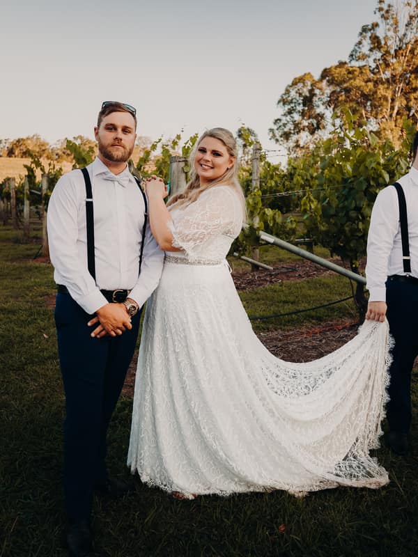 The bride Annie stands between two groomsmen at Ocean View Estates, with one groomsman holding the train of her lace wedding dress.